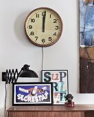 Old station clock above black, extending, retro wall lamp and framed posters on sideboard