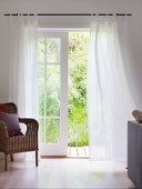 Rattansessel neben Sprossentür mit luftigem, weißem Vorhang und Blick in Garten
