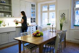 Kitchen-dining room with wooden dining table and white chairs with gingham seat cushions; woman standing at kitchen counter