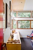 Blick auf Sideboard aus hellem Holz an Wand, im Hintergrund Fensterbänder mit Ausblick auf Bäume