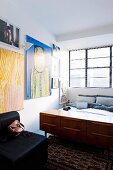 Retro Sideboard am Bettende im Schlafzimmer mit Industrie-Fenster und Kunstsammlung an der Wand