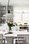 Nostalgic kitchen lamps above glass vase of roses on dining table in white, Swedish country-house kitchen