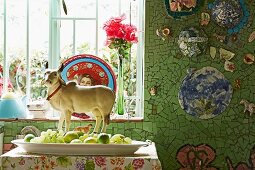 Tierfigur hinter Schale mit verschiedenen, grünen Früchten, vor Fenster, seitlich an Wand mosaikartige Fliesenstücke in Grün