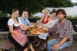 Five people sitting in a beer garden at a table with tankards and pretzels