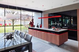 Teilweise sichtbarer Essplatz vor Kochbereich mit Küchenblock unter roter, filigraner Hängeleuchte und Einbauschrank mit schwarzer Front, seitlich Glasfassade mit Blick in Innenhof
