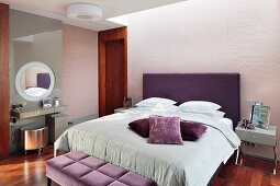 Elegantes Schlafzimmer, Doppelbett mit violett bezogenem Kopfteil, auf hellgrauer Tagesdecke Kissen in Violetttönen, gegenüber minimalistischer Schminktisch mit silberfarbenem Hocker