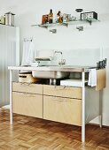 Modern kitchen sink unit with metal frame and wooden drawers below kitchen utensils on glass, wall-mounted shelf