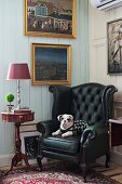 Dog on black leather armchair next to delicate, antique side table below gilt-framed pictures on white-painted wooden wall