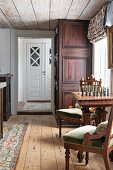 Antique upholstered chairs and chess table next to window in rustic interior with wooden floor and ceiling