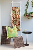 Cushion on curved rattan chair next to side table and brightly-coloured ethnic textile hanging over white-painted ladder leaning against wall