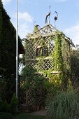 Tower-shaped lattice pavilion covered in climbing plants and decorated with bird ornaments