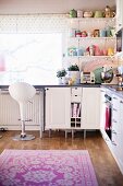 Pastel, retro crockery on open-fronted shelving, white bar stool at kitchen counter and floral rug on parquet floor
