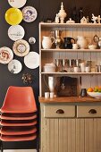 Stack of orange shell chairs below decorative plates on dark brown wall next to country-style dresser