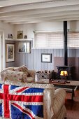 Sofa mit englischer Flagge an Rückseite und Kaminofen mit Feuer, in ländlichem Wohnraum mit holzverkleideter Wand und Holzbalkendecke