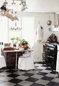 Rustikale Küche mit Schachbrettmusterboden, Stühle mit weissen Hussen am Esstisch vor Fenster, seitlich Vintage Küchenofen
