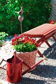 Rotes Ambiente mit Holzbank, Giesskanne und Blumentopf auf Kiesplatz im Garten