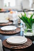 Table festively set with white plates on wicker place mats and glass vase of tulips