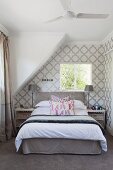 Double bed below sloping ceiling in bedroom with classic, white and grey patterned wallpaper