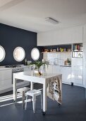 Table on castors and vintage stool in white fitted kitchen with black mosaic floor, black accent wall and porthole windows