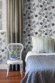 White wicker chair next to bed with patterned bedspread and wallpaper with white and blue, stylised floral pattern in bedroom