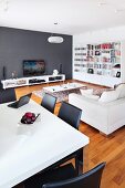 White dining table and black chairs in front of lounge area with sofa and low, white sideboard against dark grey wall