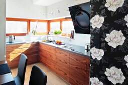 A modern fitted kitchen with wooden cupboards and windows all the way round with a piece of black and white floral wallpaper in the foreground