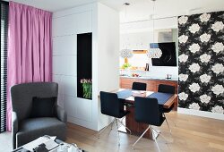 A view from a seating area looking towards a dining table with crystal pendant lamps and black and white floral wallpaper and the kitchen in the background behind a room divider cupboard