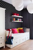 Coloured cushions on a sofa bed with drawers against a wall hung with black-and-white spotted wallpaper