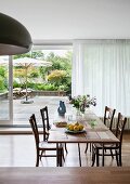 Dining table with wooden top and old bentwood chairs in front of glass wall with half-closed curtain showing view of summery terrace