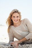 A young blonde woman lying on a beach wearing a beige knitted jumper