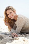 A young blonde woman lying on a beach wearing a beige knitted jumper