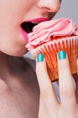 A young woman biting into a pink cupcake, face cropped