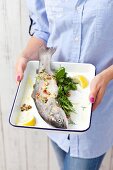 A woman holding fresh trout, herbs and lemons on a baking tray
