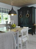 Schlichter Esstisch und weiße Küchenstühle unter Vintage Hängeleuchte, im Hintergrund dunkel lackierter Bauernschrank mit Kränzen an Türen