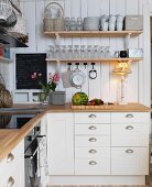 weiße Küche im Landhausstil, Tischleuchte auf Küchenzeile, unter Wandkonsolen mit Gläsern und Bechern an Holzwand aufgehängt