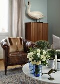 Candlestick with frog-shaped base and vase of flowers on coffee table, old leather armchair and stuffed mute swan on cabinet in background