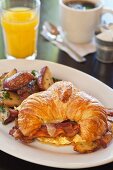 A croissant with scrambled eggs, bacon and cheese served with a side of sauteed potatoes, orange juice and coffee