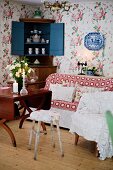 Various chairs, wooden table, sofa, crockery in open corner cabinet and floral wallpaper