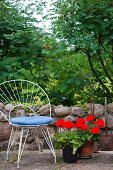 Weisser, filigraner Metallstuhl mit blauem Sitzkissen, neben roten Geranien auf Natursteinboden, vor niederiger Gartenmauer mit Blick in Garten