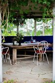 Rustikaler Holztisch und weiße, filigrane Metallstühle auf Natursteinboden in Loggia