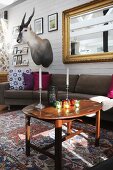 Candles on tray table in front of sofa with scatter cushions below gilt-framed mirror and hunting trophy on wall