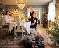 Kleines Kind und Hund auf Teppich neben Weihnachtsbaum, im Hintergrund Familie an Festtafel in traditionellem Esszimmer