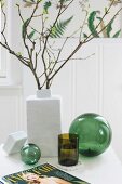 Blätterzweige in weisser Keramikvase und grünes Trinkglas zwischen verschiedenen grünen Glaskugeln auf weißem Tisch