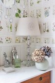 Tischleuchte mit weißem Schirm und Hortensien in Kugelvase auf Kommode, an Wand Tapete mit botanischem Muster