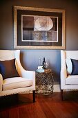Delicate wire mesh side table flanked by elegant armchairs with white upholstery below framed picture on light grey wall