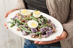 A woman holding a plate of purple potato salad with green beans, egg, tuna fish and mayonnaise