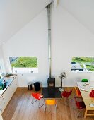 View down into interior with kitchen counter, dining area with colourful shell chairs and wood-burning stove between windows