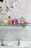 Ornate mirror frame, floral wallpaper and glass shelf of romantic knickknacks above vintage tap fittings