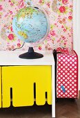 Globe lamp on white, retro sideboard with yellow-painted cupboard doors and red and white polka-dot suitcase to one side