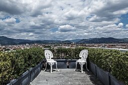 Zwei weiße Vintage Metallstühle im Rokoko Stil in den Ecken der bepflanzten Dachterrasse positioniert mit herrlichem Panoramablick